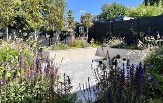 Raised sleeper beds, made of light grey porcelain tiles in paved garden. After image showing borders blooming and overflowing with flowers and plants of all different shapes and colours. Designed and landscaped by the Essex Garden Co.