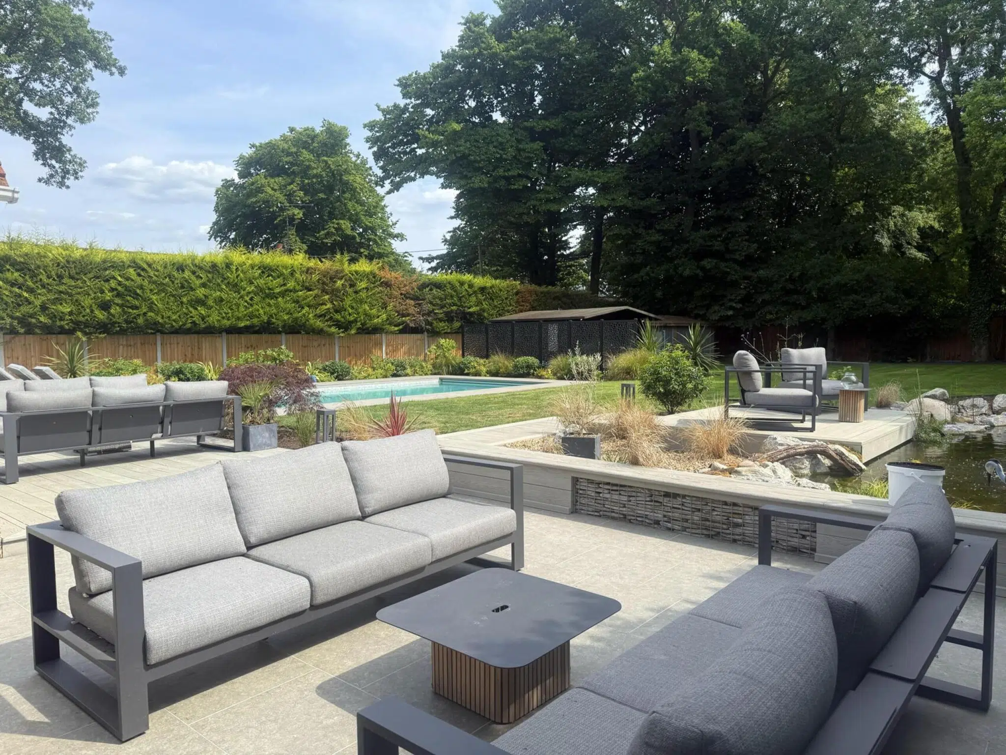 Blackmore, Essex garden transformation. Low maintenance garden with lots of lounging areas. Features include porcelain paving, composite decking, pond, outdoor kitchen, planting, lighting, swimming pool. Professional landscaping by award winning gardening team, The Essex Garden Co.